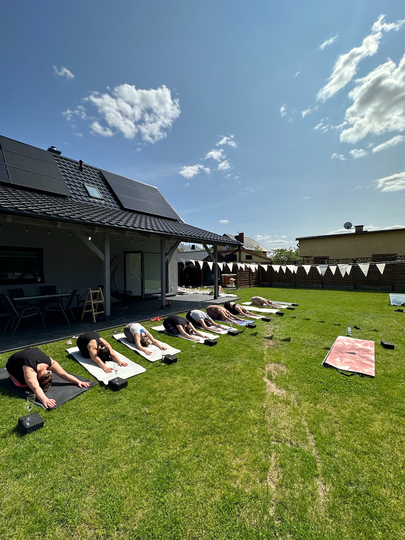 Grupa osób wykonuje pozycję dziecka podczas zajęć jogi na trawie przed domem z panelami słonecznymi, białe chorągiewki dekoracyjne zawieszone w tle, szklanki z napojami obok mat.