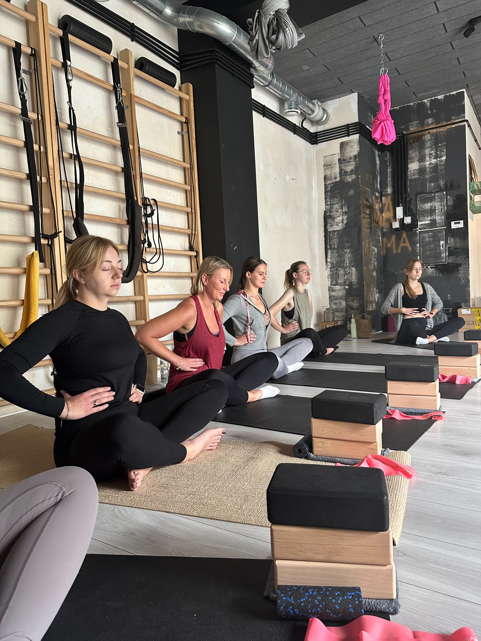 Grupa kobiet siedzi w rzędzie na matach do jogi, wykonując ćwiczenie z rękami na brzuchu, w tle drabinki gimnastyczne i podwieszana huśtawka do aerial jogi.