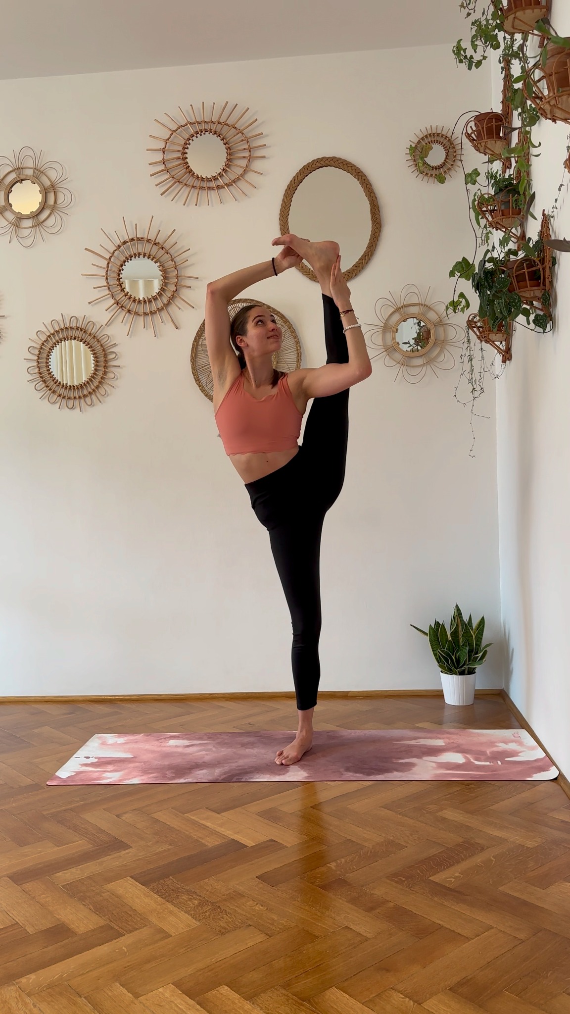 Kobieta w różowym topie i czarnych legginsach wykonuje zaawansowaną pozycję jogi na różowej macie w jasnym pokoju z drewnianą podłogą w jodełkę, ozdobionym lustrami i roślinami.