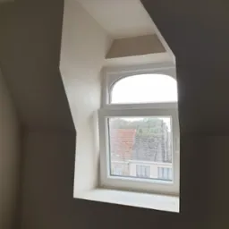 Poddasze po pracach wykończeniowych, widoczne okno z kroplami deszczu i świeżo położone tynki na skosach ścian.