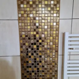 Fragment ściany łazienki z dekoracyjną wstawką z mozaiki w odcieniach złota i brązu, obok biały grzejnik drabinkowy.