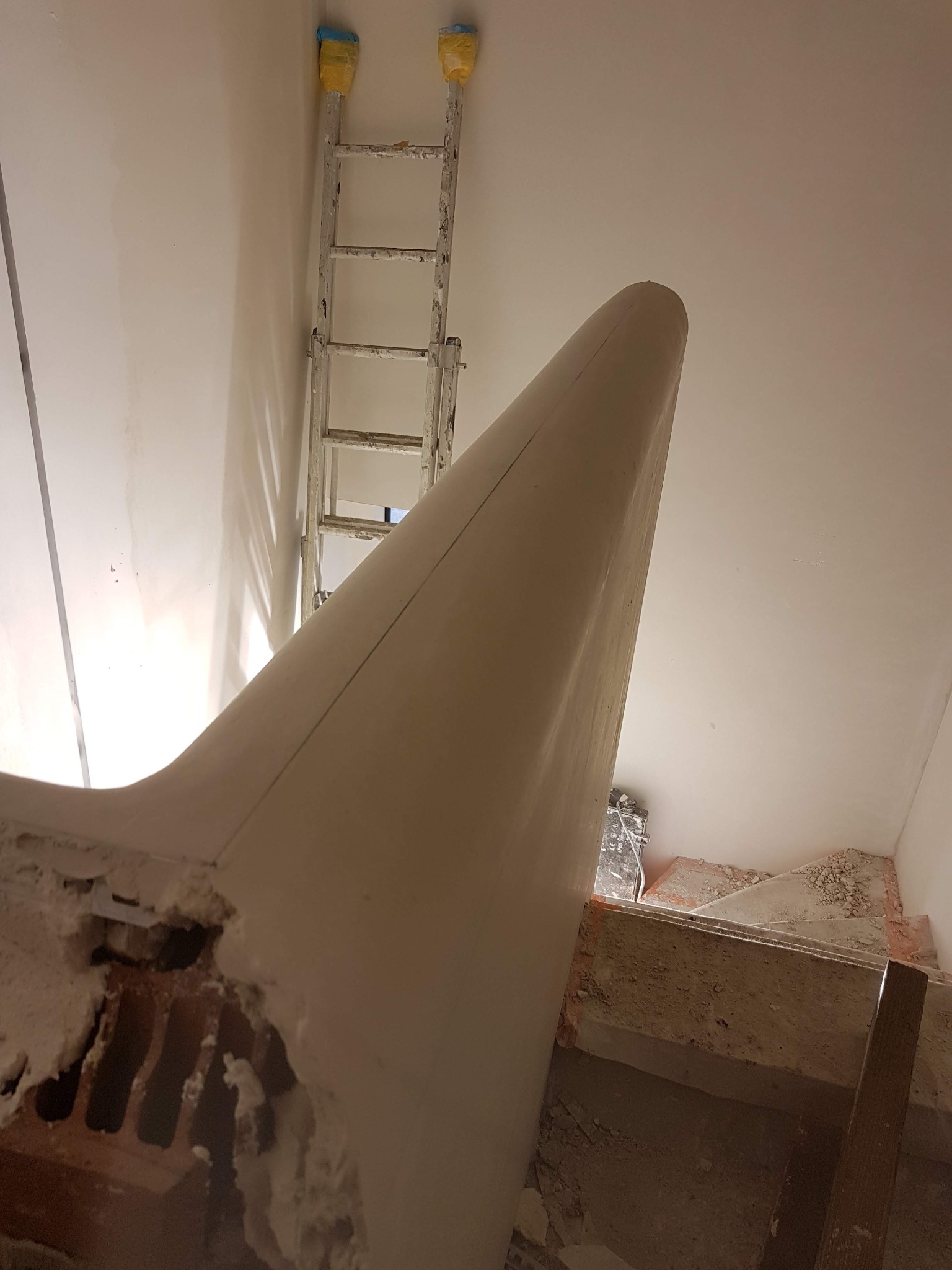 Wnętrze w trakcie prac wykończeniowych: widok z dołu na uformowany element architektoniczny na suficie, częściowo pokryty białą masą, z widocznym fragmentem drabiny opierającej się o ścianę...