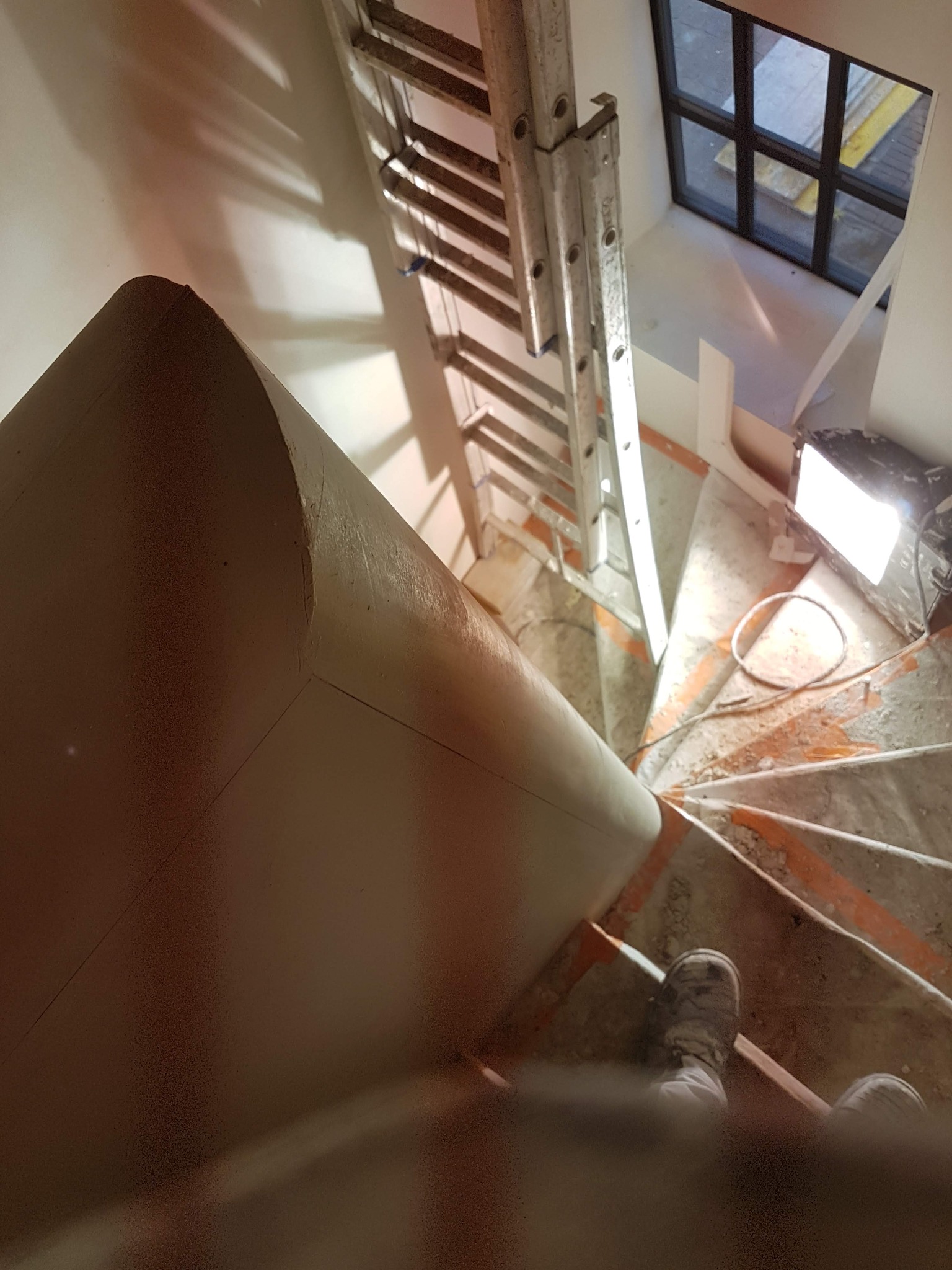 Widok z góry na spiralne schody w trakcie prac wykończeniowych, widoczna drabina oparta o ścianę, lampa halogenowa oświetlająca przestrzeń, zabezpieczone taśmą malarską krawędzie stopni i fragment...