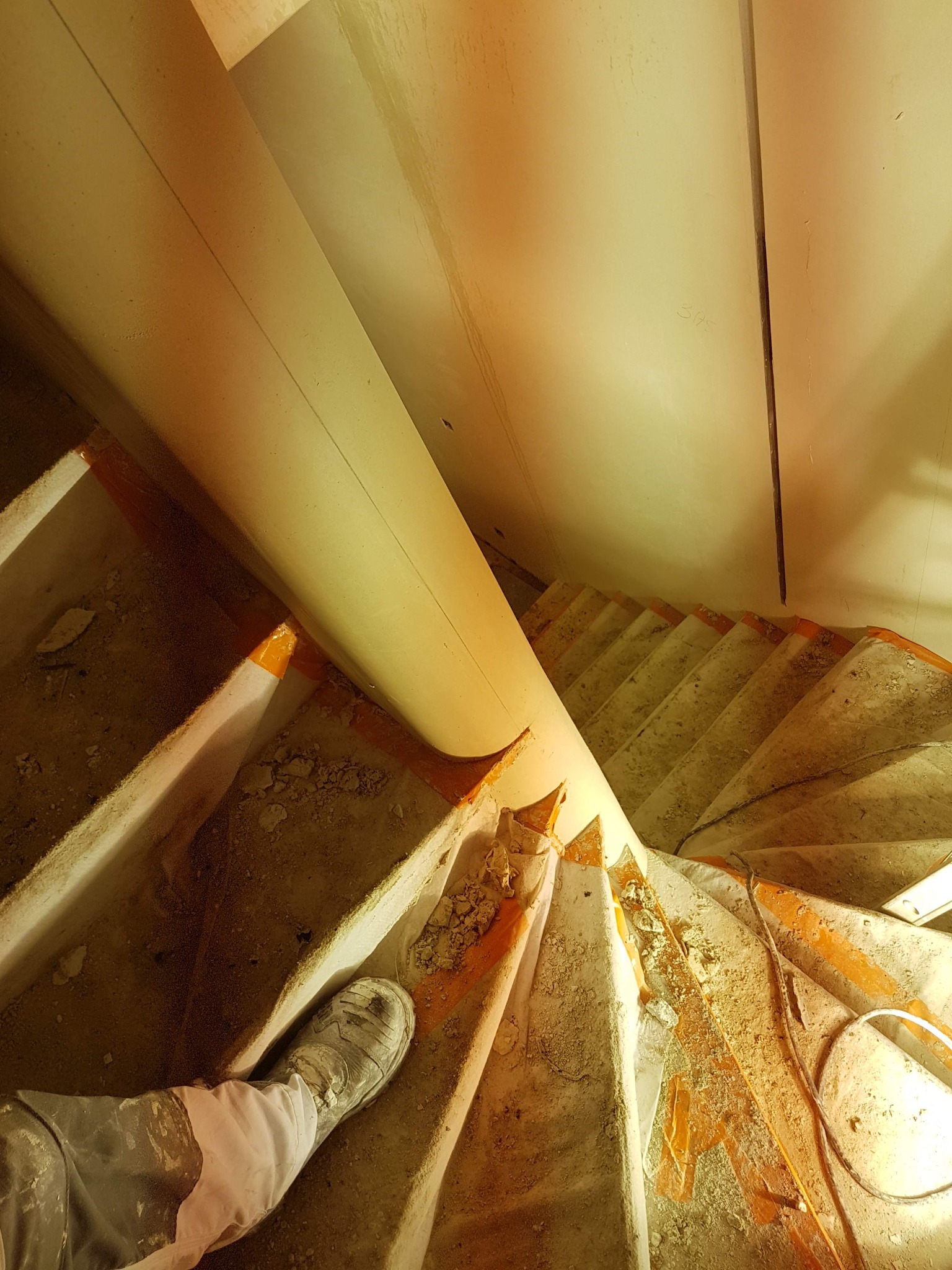 Wewnętrzne schody spiralne w trakcie prac wykończeniowych, zabezpieczone pomarańczową taśmą, z widoczną nogą w zabrudzonym bucie roboczym i częściowo otynkowaną ścianą.