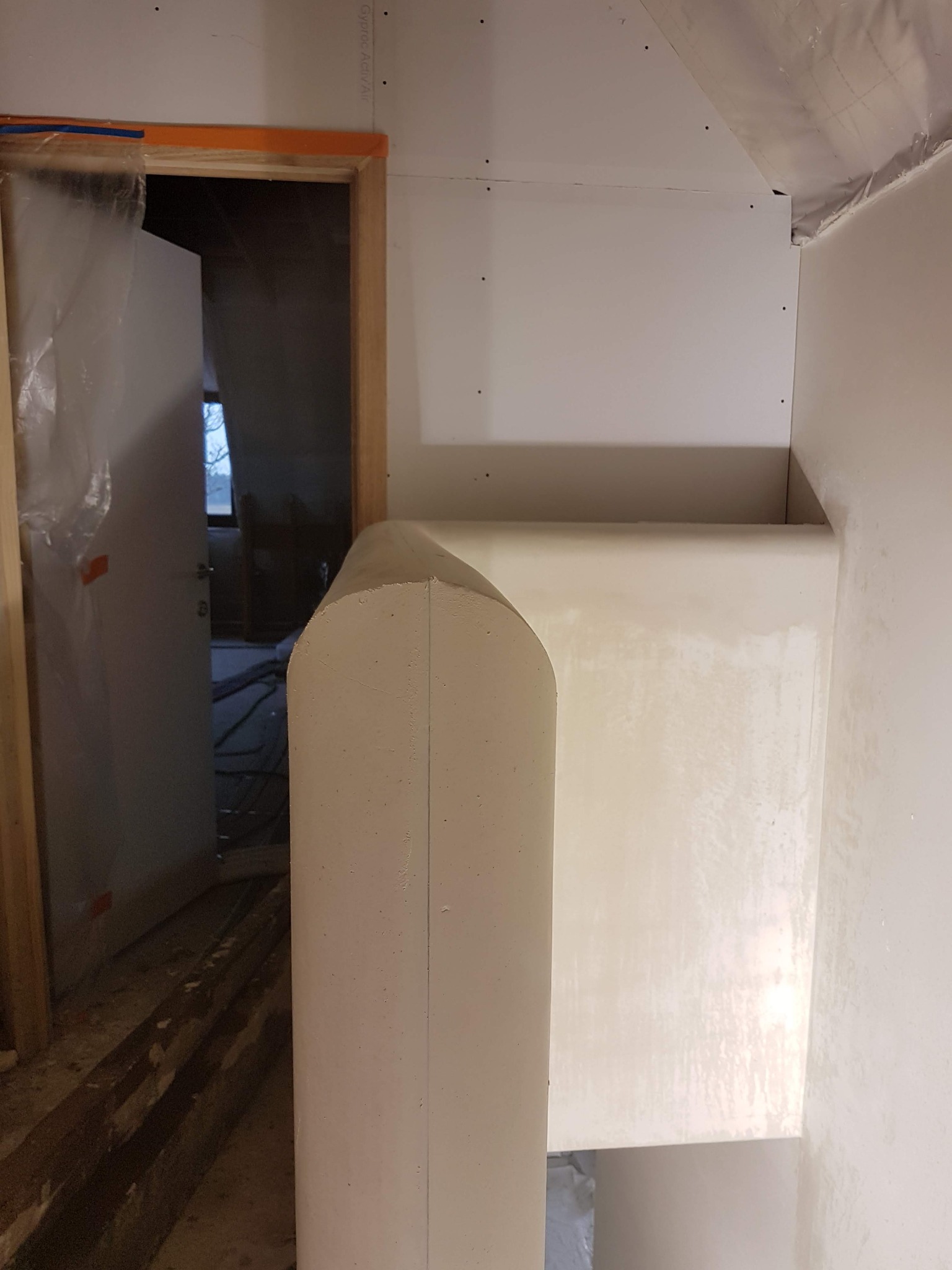 Wnętrze w trakcie prac wykończeniowych: zaokrąglona kolumna po obróbce, widoczne płyty gipsowo-kartonowe na ścianach i przygotowane do dalszych prac wykończeniowych futryny drzwiowe.