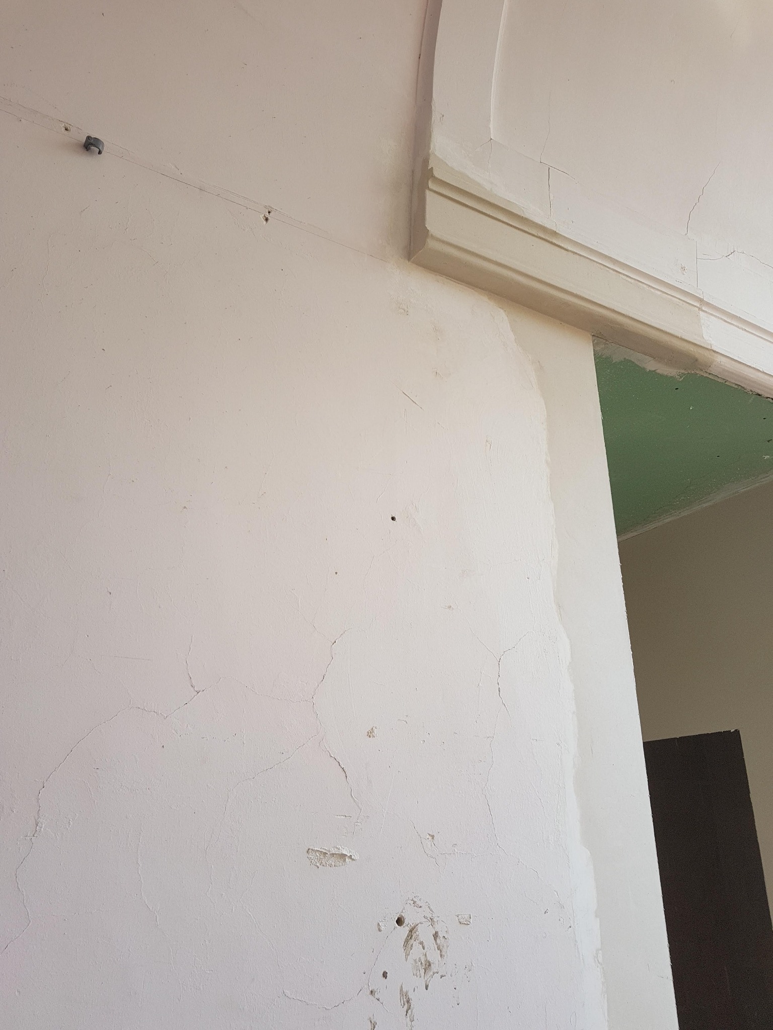 Fragment ściany z widocznymi pęknięciami i śladami po starym szpachlowaniu, częściowo pokrytej nową warstwą gładzi, z widoczną listwą dekoracyjną przy suficie również częściowo wyszpachlowaną.