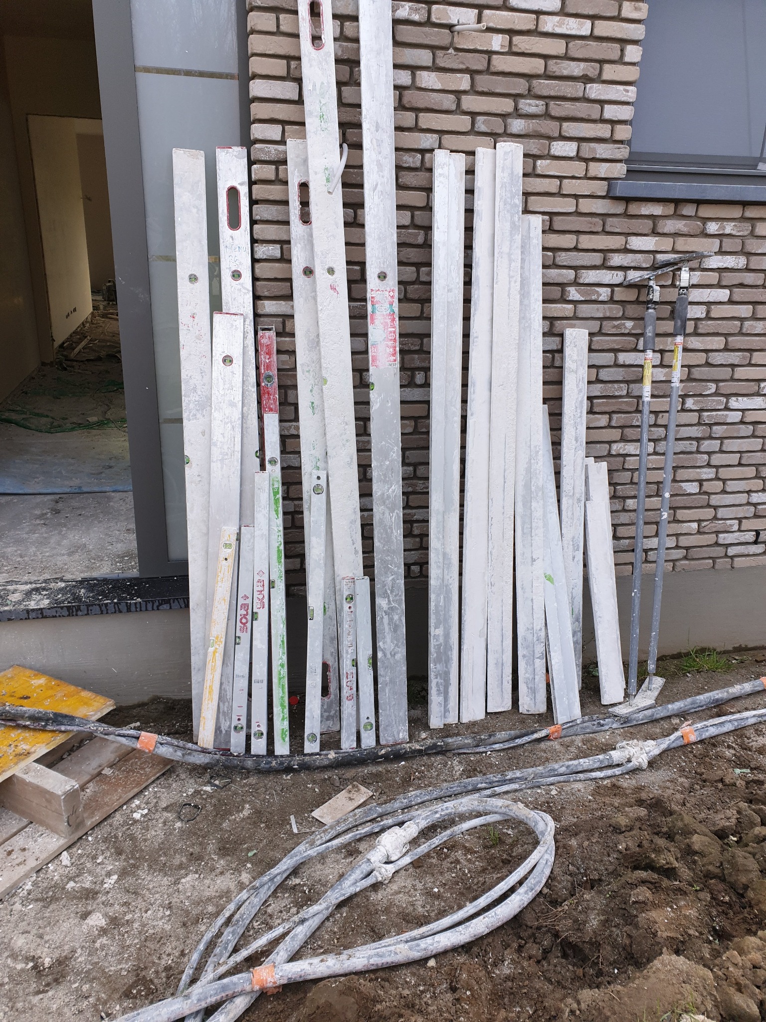 Stojące pionowo poziomice różnych długości i profile tynkarskie opierają się o ceglaną ścianę domu w trakcie prac wykończeniowych; widoczne kable i wejście do budynku.