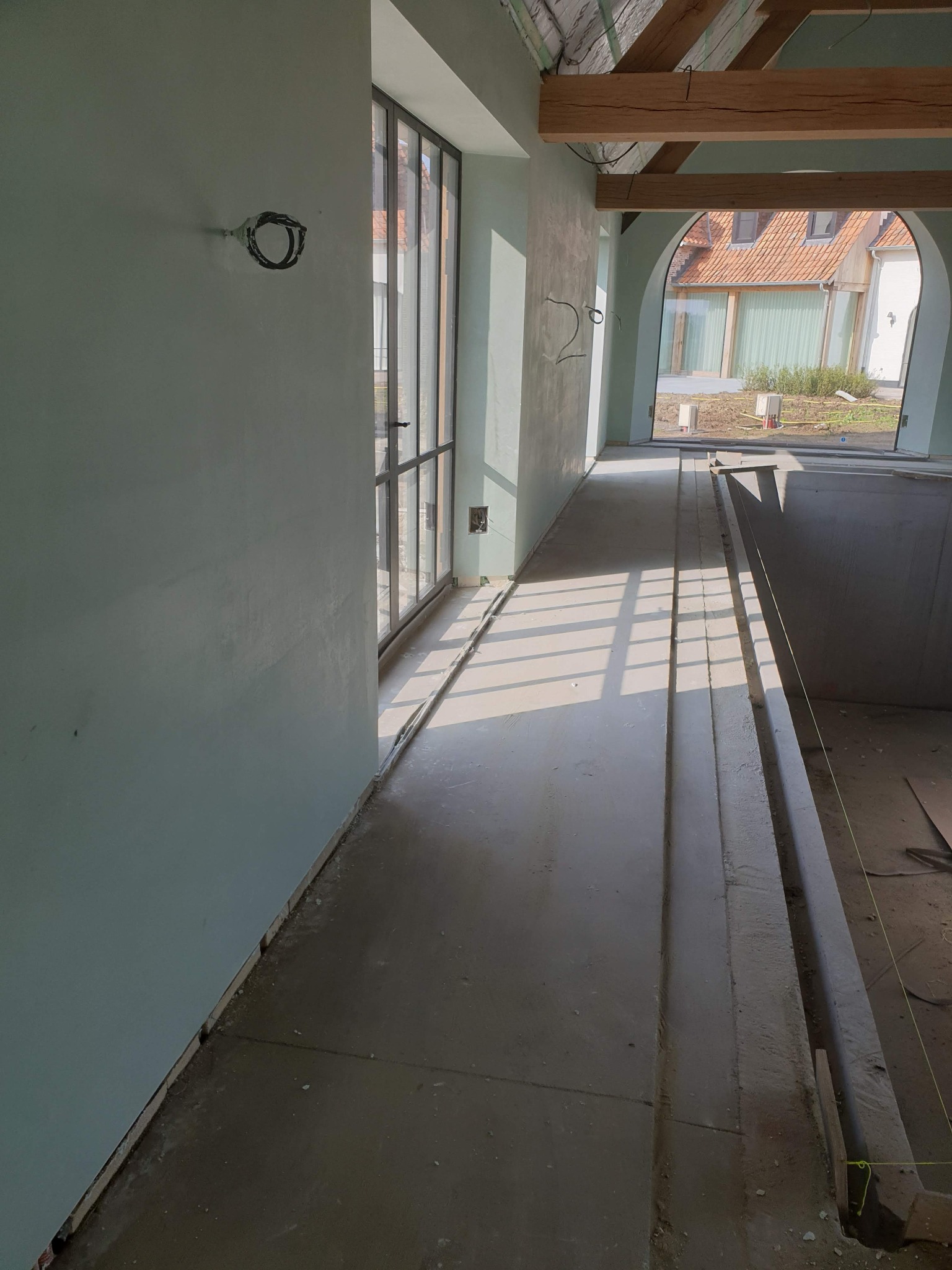 Wnętrze w trakcie prac wykończeniowych: ściany pokryte świeżym, jasnym tynkiem, widoczne kable elektryczne, duże okno z widokiem na budynek z czerwoną dachówką, drewniane elementy konstrukcyjne...