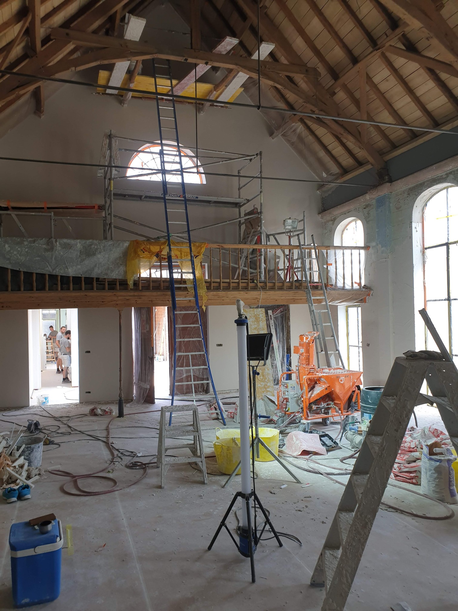 Przestronne wnętrze w trakcie prac wykończeniowych: rusztowania, drabiny, agregat tynkarski, ściany przygotowane pod malowanie, widoczne drewniane elementy konstrukcyjne dachu i antresoli.