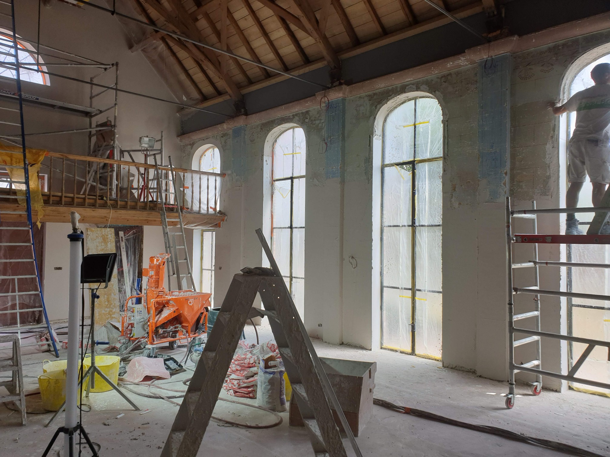 Wnętrze w trakcie tynkowania maszynowego: widoczne rusztowania, drabiny, maszyna do tynkowania, worki z zaprawą, okna zabezpieczone folią oraz tynkarz pracujący przy oknie na rusztowaniu.