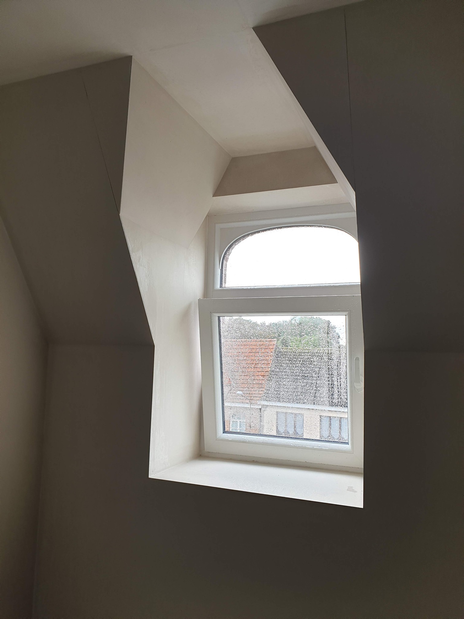 Poddasze po pracach wykończeniowych, widoczne okno z kroplami deszczu i świeżo położone tynki na skosach ścian.