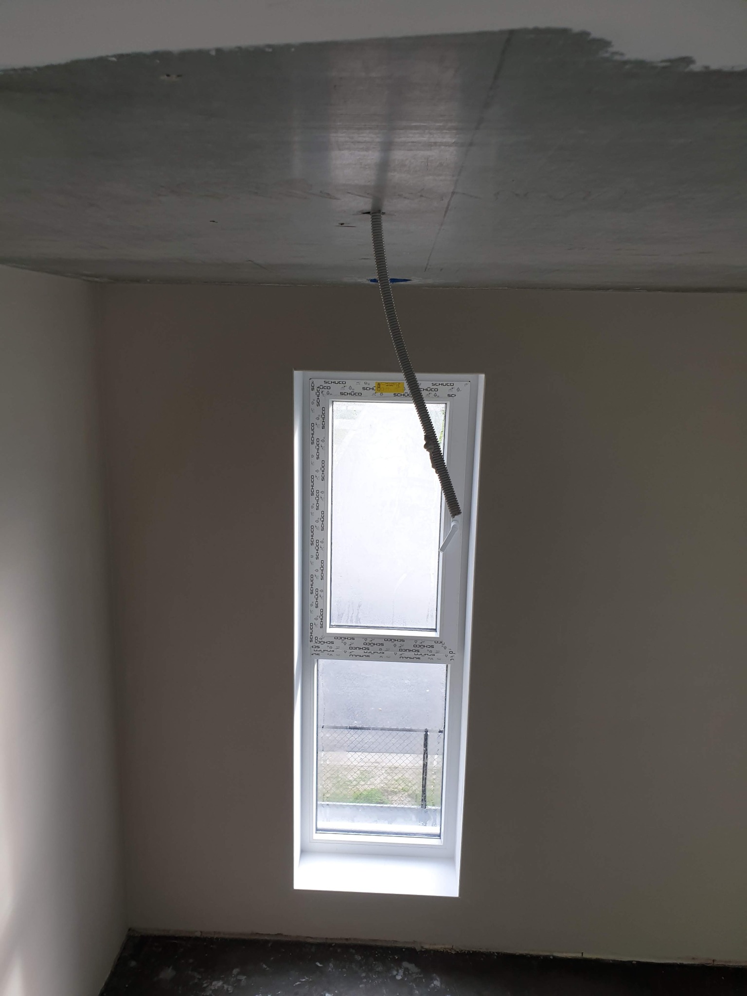 Surowy betonowy sufit w trakcie prac wykończeniowych, widoczne szpachlowanie przy krawędziach, z wiszącą rurą instalacyjną obok okna PCV z widokiem na zewnątrz.