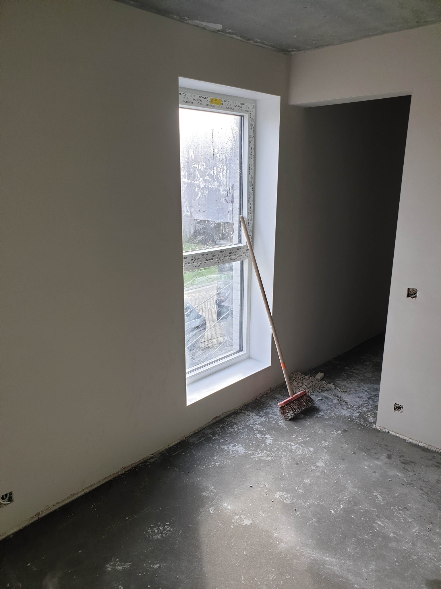 Pokój w stanie deweloperskim po pracach, ściany pokryte gładzią, widoczny fragment okna z naklejkami producenta, na podłodze resztki gruzu i śmieci, oparta o ścianę czerwona zmiotka.
