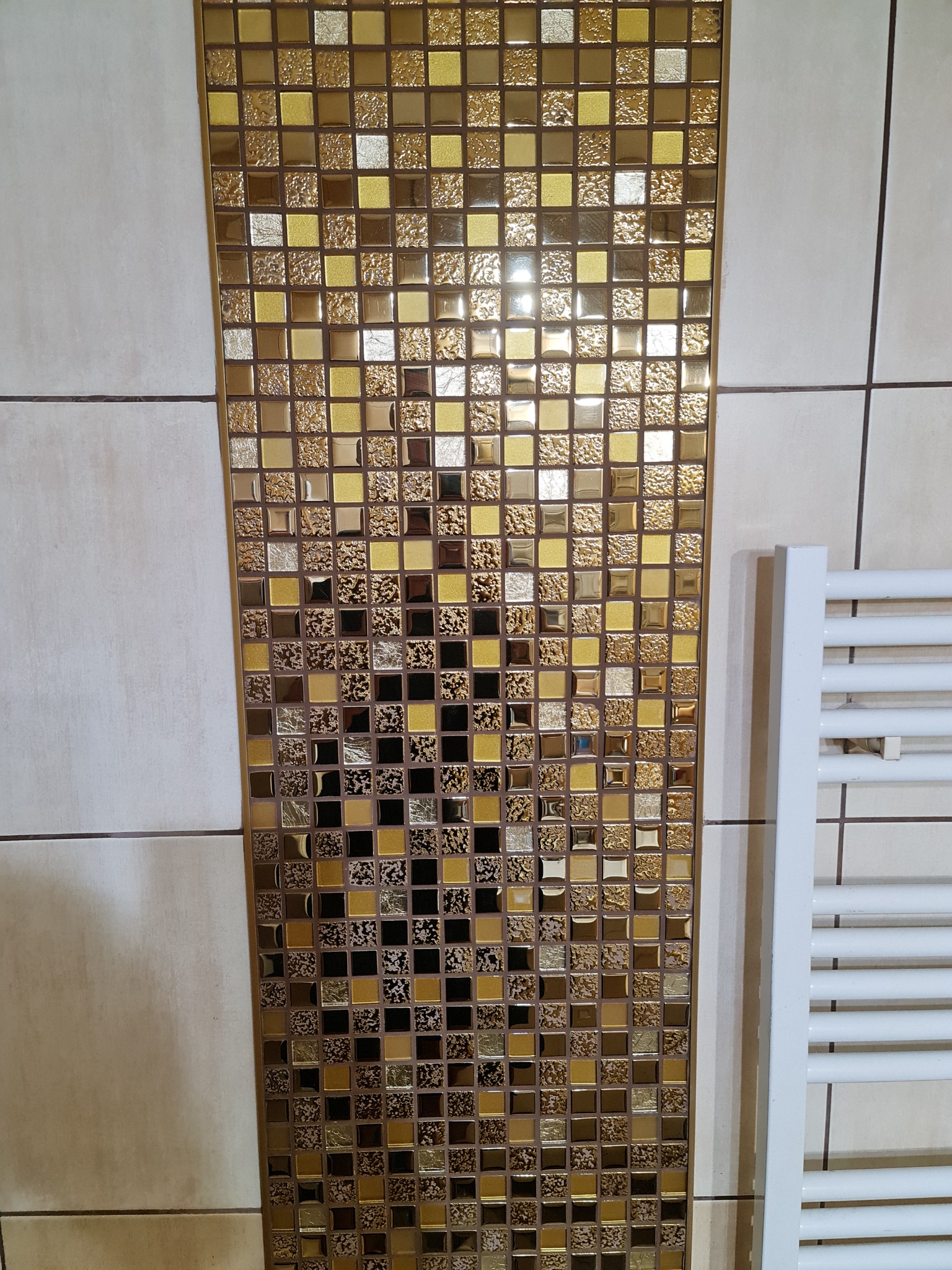 Fragment ściany łazienki z dekoracyjną wstawką z mozaiki w odcieniach złota i brązu, obok biały grzejnik drabinkowy.