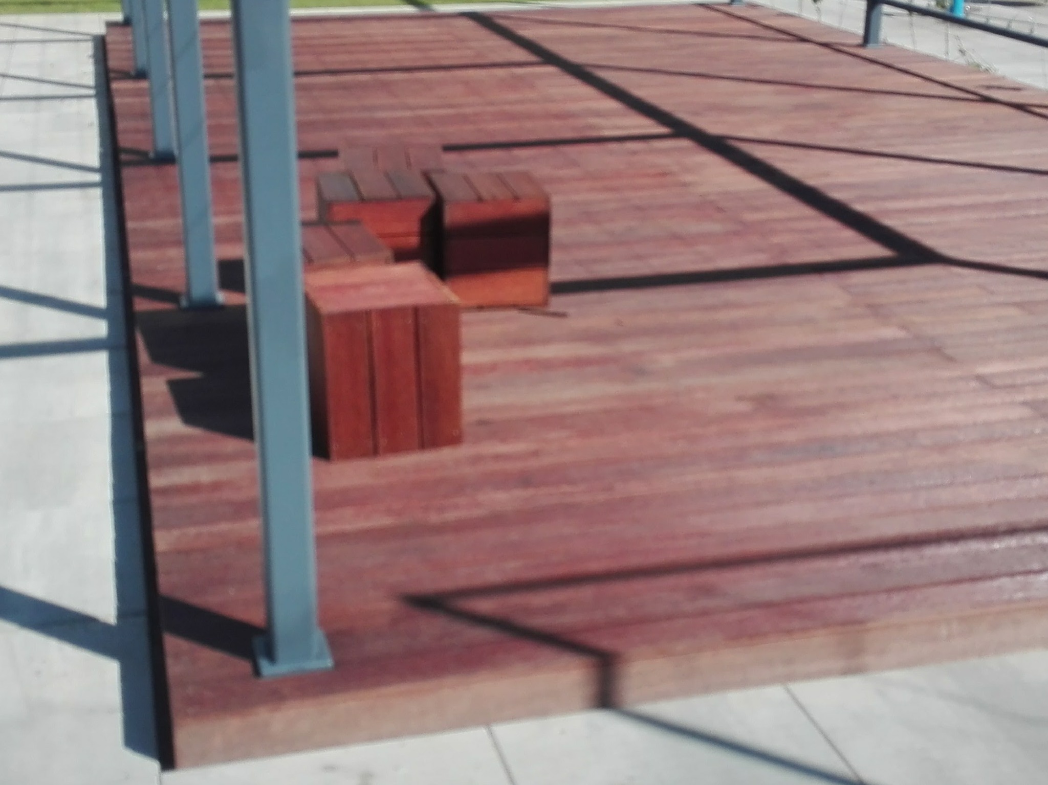 Drewniany taras z trzema drewnianymi siedziskami w kształcie kostek, oświetlony promieniami słońca padającymi przez konstrukcję z metalowymi słupkami.