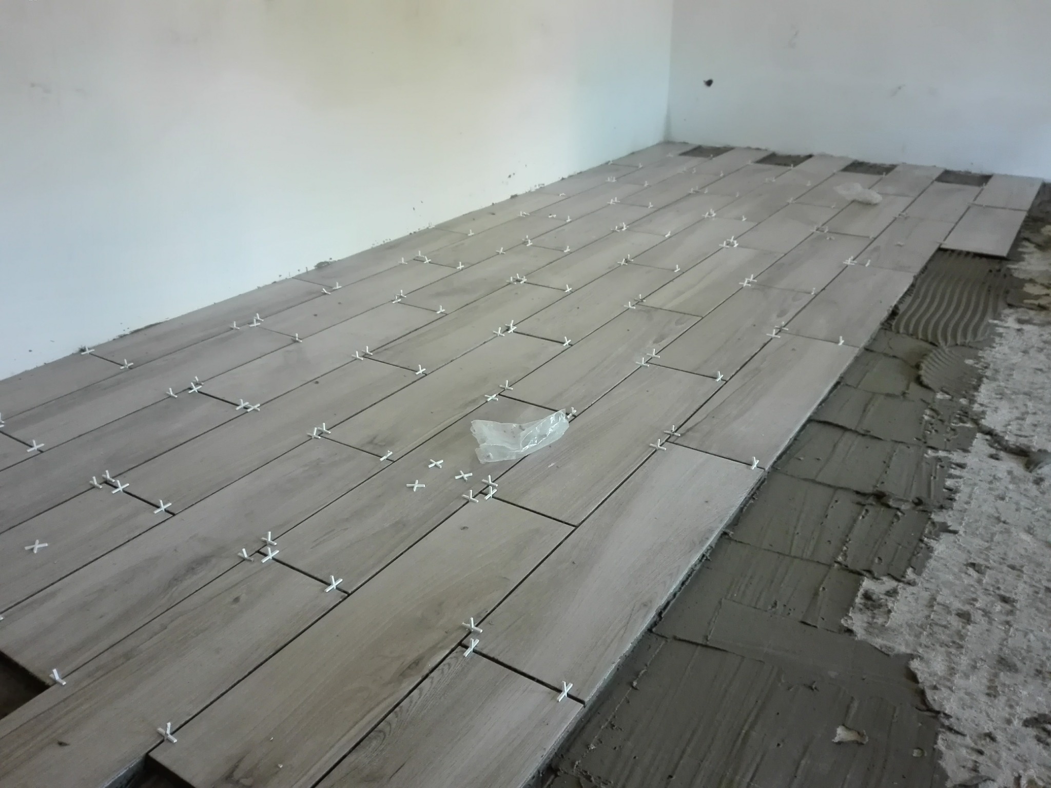 Układanie podłogi z płytek imitujących drewno w jasnym odcieniu, z użyciem białych krzyżyków dystansowych, widoczny fragment nałożonego kleju na podłożu.