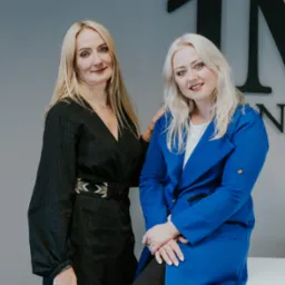 Dwie uśmiechnięte blondynki w eleganckich strojach pozują w biurze firmy w Gnieźnie, widoczne logo na ścianie w tle.