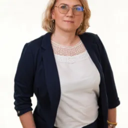 Portret kobiety w okularach, białej bluzce z koronkowym wykończeniem i granatowej marynarce, pozującej na jasnym tle.