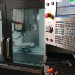Wnętrze maszyny CNC z widocznym uchwytem narzędziowym i obrabianym elementem. Na pierwszym planie panel sterowania z przyciskiem awaryjnym i ekranem LCD. Zabrudzona szyba ochronna maszyny.