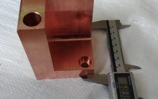 Miedziany blok z dwoma otworami, mierzony suwmiarką elektroniczną wskazującą 68.89 mm. Widoczne ślady obróbki i niedoskonałości powierzchni.