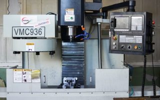 Pionowe centrum obróbcze VMC936 z panelem sterowania, widoczne ostrzeżenia BHP i częściowo widoczny detal w obszarze roboczym.