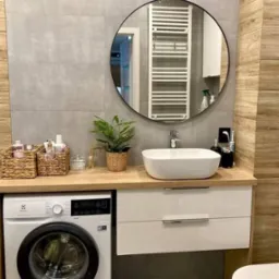 Nowoczesna łazienka z pralką Electrolux, białą umywalką nablatową, okrągłym lustrem z czarną ramą i grzejnikiem drabinkowym.