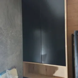 Ciemne, minimalistyczne szafki łazienkowe z otwartą półką z jasnego drewna, na tle szarej ściany z płytek i fragmentu drewnianej okładziny.