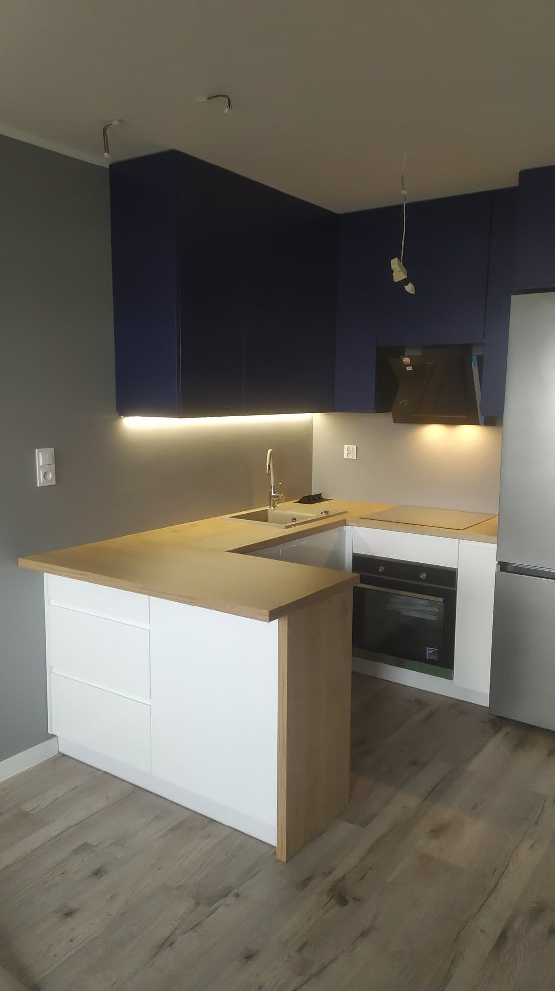Nowoczesna kuchnia z granatowymi szafkami wiszącymi, drewnianym blatem i białymi szafkami dolnymi, widoczne niedokończone podłączenia elektryczne na suficie.