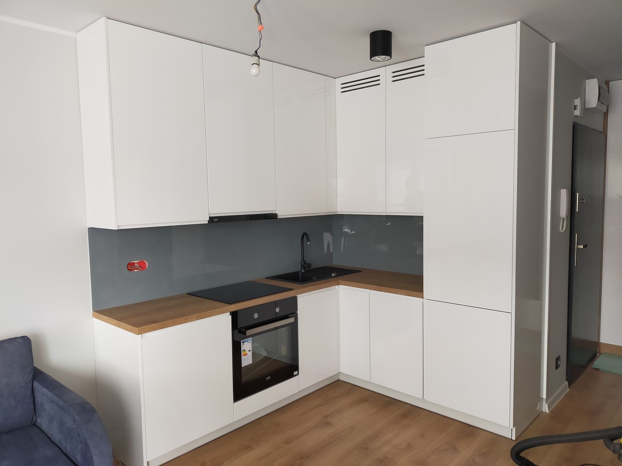 Nowoczesna kuchnia w kształcie litery L z białymi, błyszczącymi frontami, drewnianym blatem, szarym panelem nad blatem i czarną armaturą, widok na fragment salonu z niebieskim fotelem.