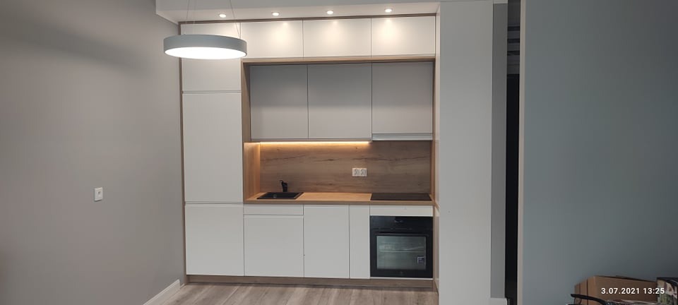 Nowoczesna, minimalistyczna zabudowa kuchenna z białymi frontami i drewnianym blatem, oświetlona światłem LED pod szafkami wiszącymi, z czarnym zlewem i płytą indukcyjną.