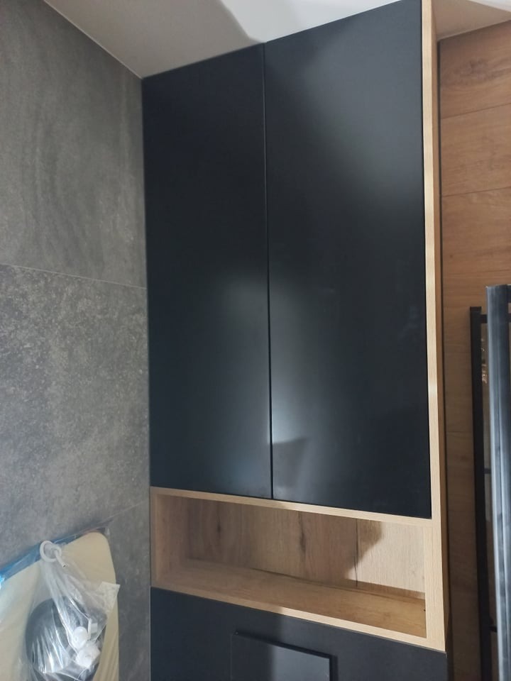 Ciemne, minimalistyczne szafki łazienkowe z otwartą półką z jasnego drewna, na tle szarej ściany z płytek i fragmentu drewnianej okładziny.