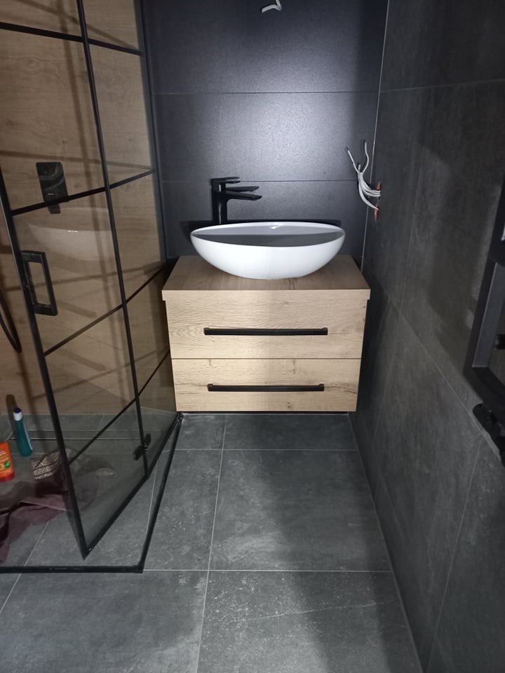 Nowoczesna łazienka z umywalką nablatową w kształcie misy, czarną armaturą i szafką z drewnopodobnym frontem, widoczna kabina prysznicowa z czarnymi profilami.