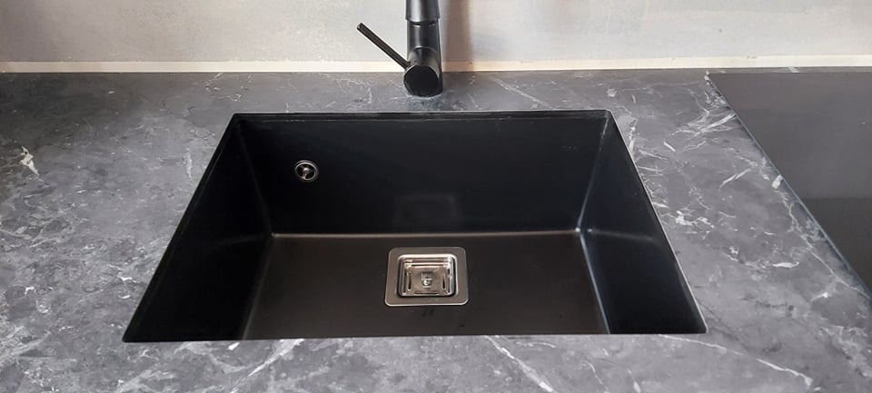 Czarny, prostokątny zlew granitowy z chromowanym odpływem i czarną baterią, osadzony w blacie imitującym marmur.