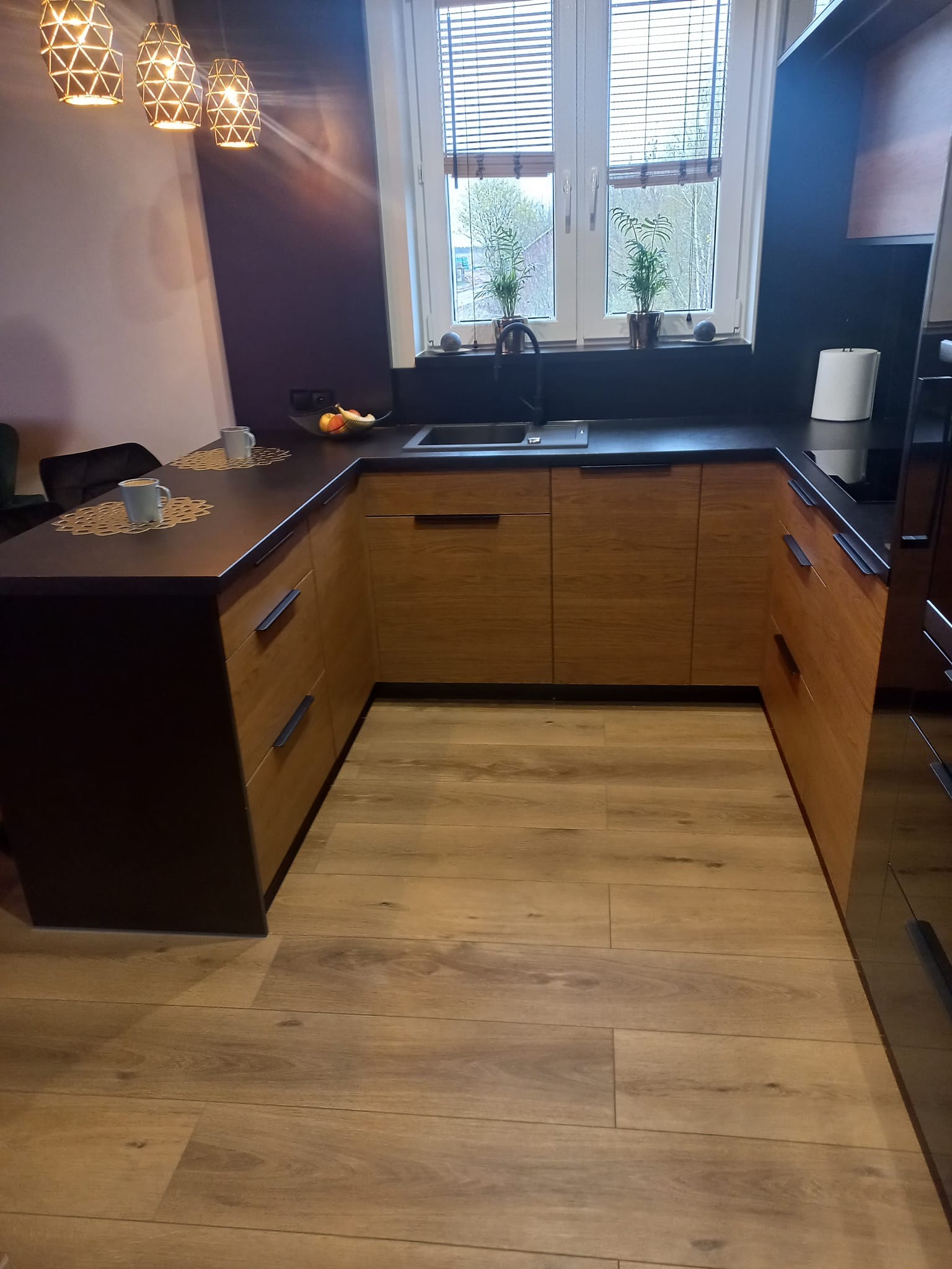 Nowoczesna kuchnia w kształcie litery U z drewnianymi frontami szafek, czarnym blatem i podłogą z paneli imitujących drewno. Na blacie widoczne filiżanki, owoce i ozdobne podkładki. Nad blatem...