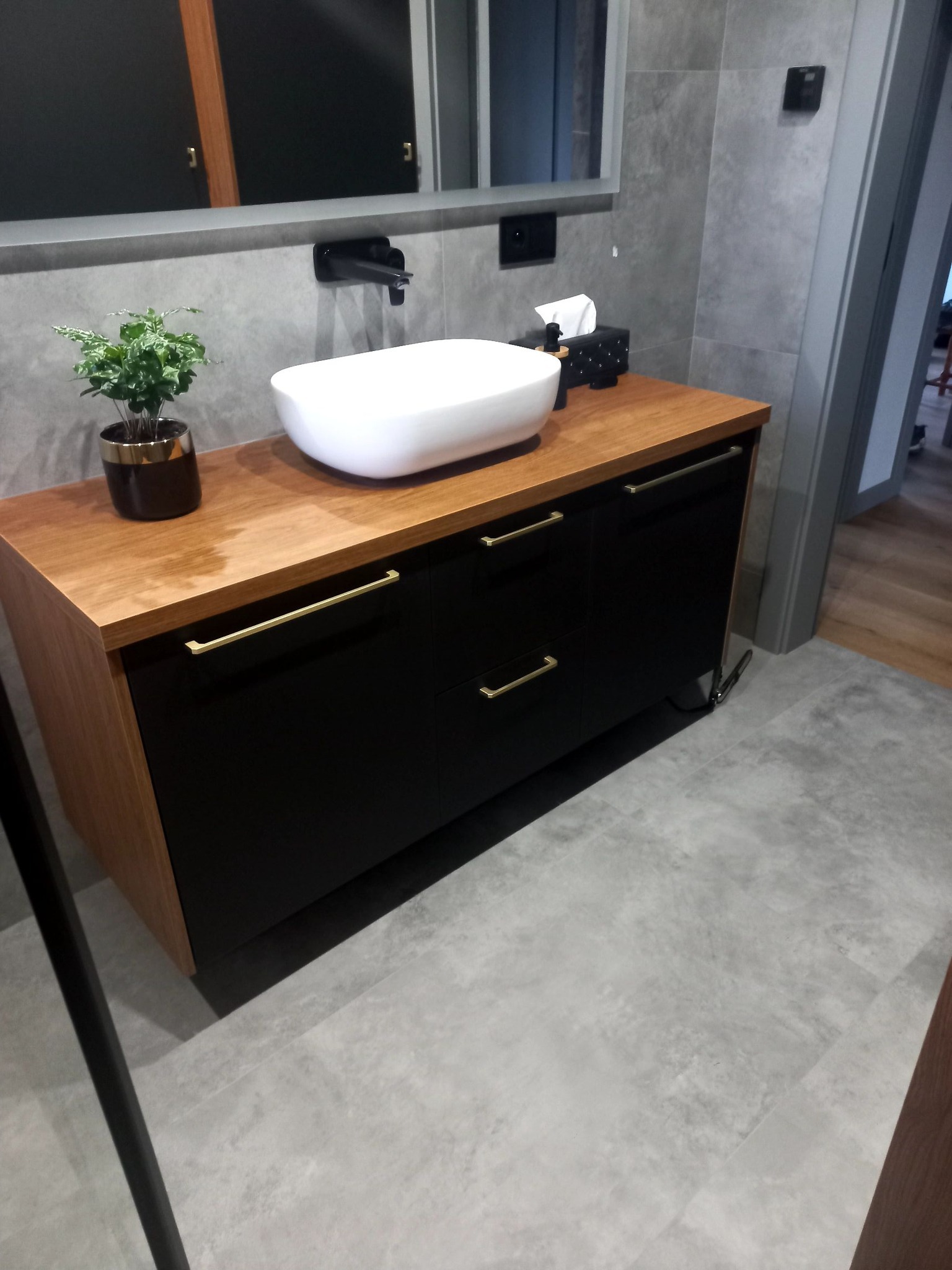 Nowoczesna łazienka z drewnianą szafką pod umywalką, czarnymi frontami, złotymi uchwytami i owalną białą umywalką nablatową, uzupełniona o czarną armaturę i dekoracyjną roślinkę w złotym wazonie.