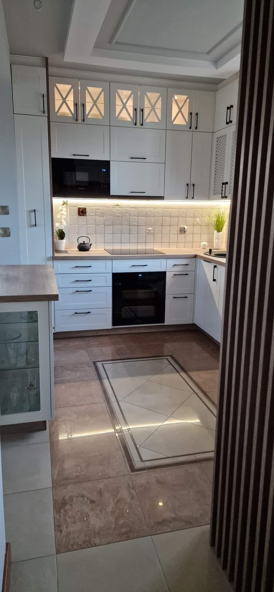 Nowoczesna, jasna kuchnia z białymi szafkami z podświetleniem, czarnymi uchwytami i sprzętami, oraz brązowymi płytkami podłogowymi z dekoracyjnym wzorem.