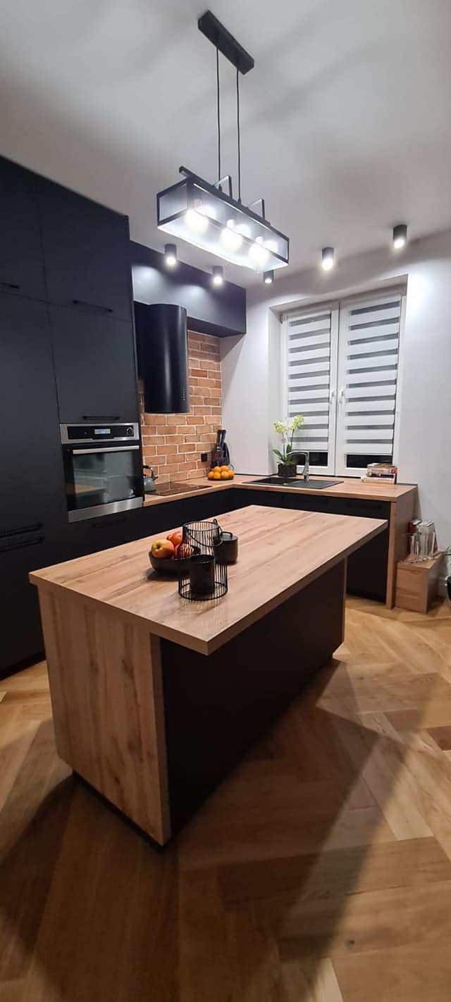 Nowoczesna kuchnia z czarnymi szafkami, drewnianym blatem wyspy, ceglaną ścianą i parkietem w jodełkę, oświetlona wiszącą lampą.