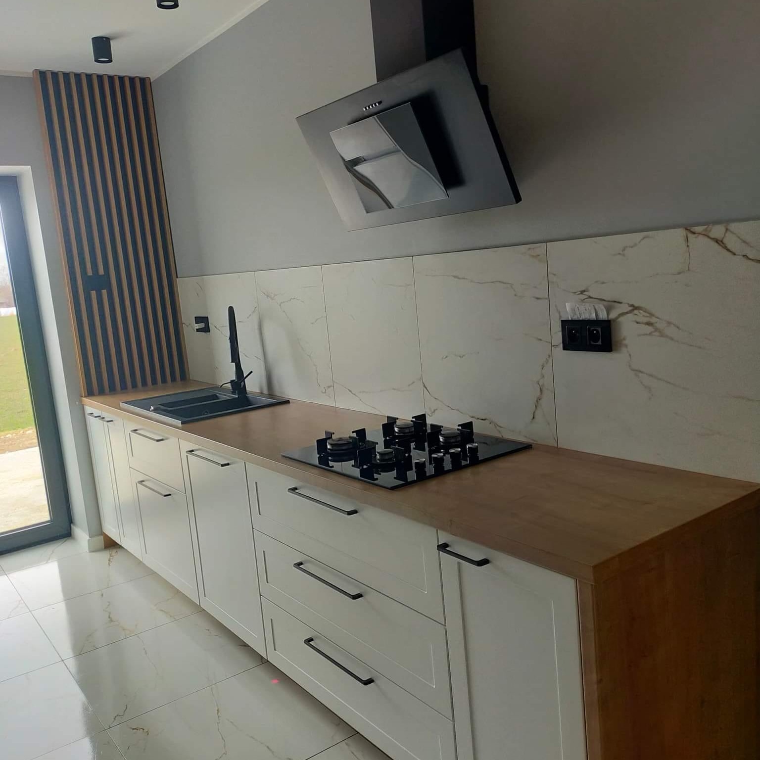 Nowoczesna kuchnia z białymi szafkami, drewnianym blatem i czarnymi uchwytami, płyta gazowa i okap w kolorze czarnym, marmurowe płytki na ścianie, lamelki dekoracyjne na ścianie obok drzwi.