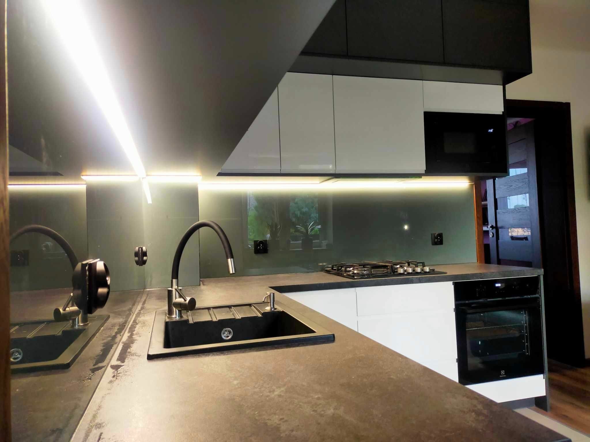 Nowoczesna kuchnia z czarnym zlewem i baterią, szarym blatem, białymi szafkami dolnymi i górnymi w kolorze czarnym i białym, oraz szklanym panelem nad blatem, oświetlona listwami LED pod szafkami.