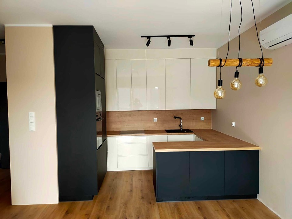 Nowoczesna kuchnia w odcieniach bieli, czerni i drewna, z wiszącą lampą z drewnianym elementem i żarówkami Edisona, oświetlona światłem punktowym, z drewnianą podłogą i klimatyzacją na ścianie.