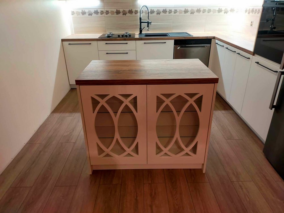 Biała kuchnia z wyspą, drewniany blat, dekoracyjne wycięcia na frontach wyspy, płytki z motywem liści na ścianie.