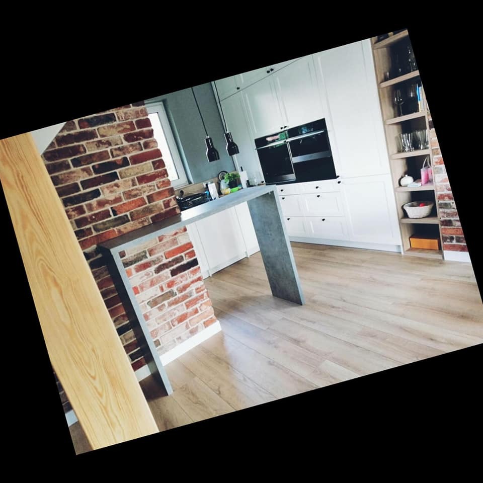 Nowoczesna biała kuchnia z ceglanym barem i drewnianą podłogą, oświetlona wiszącymi lampami, z widocznymi półkami i sprzętem AGD.