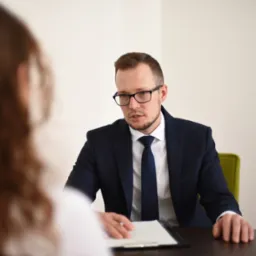 Mężczyzna w garniturze i okularach siedzi za biurkiem, prowadząc rozmowę z osobą siedzącą naprzeciwko, której widać jedynie fragment włosów i ramienia. Na biurku leży podkładka z kartkami.