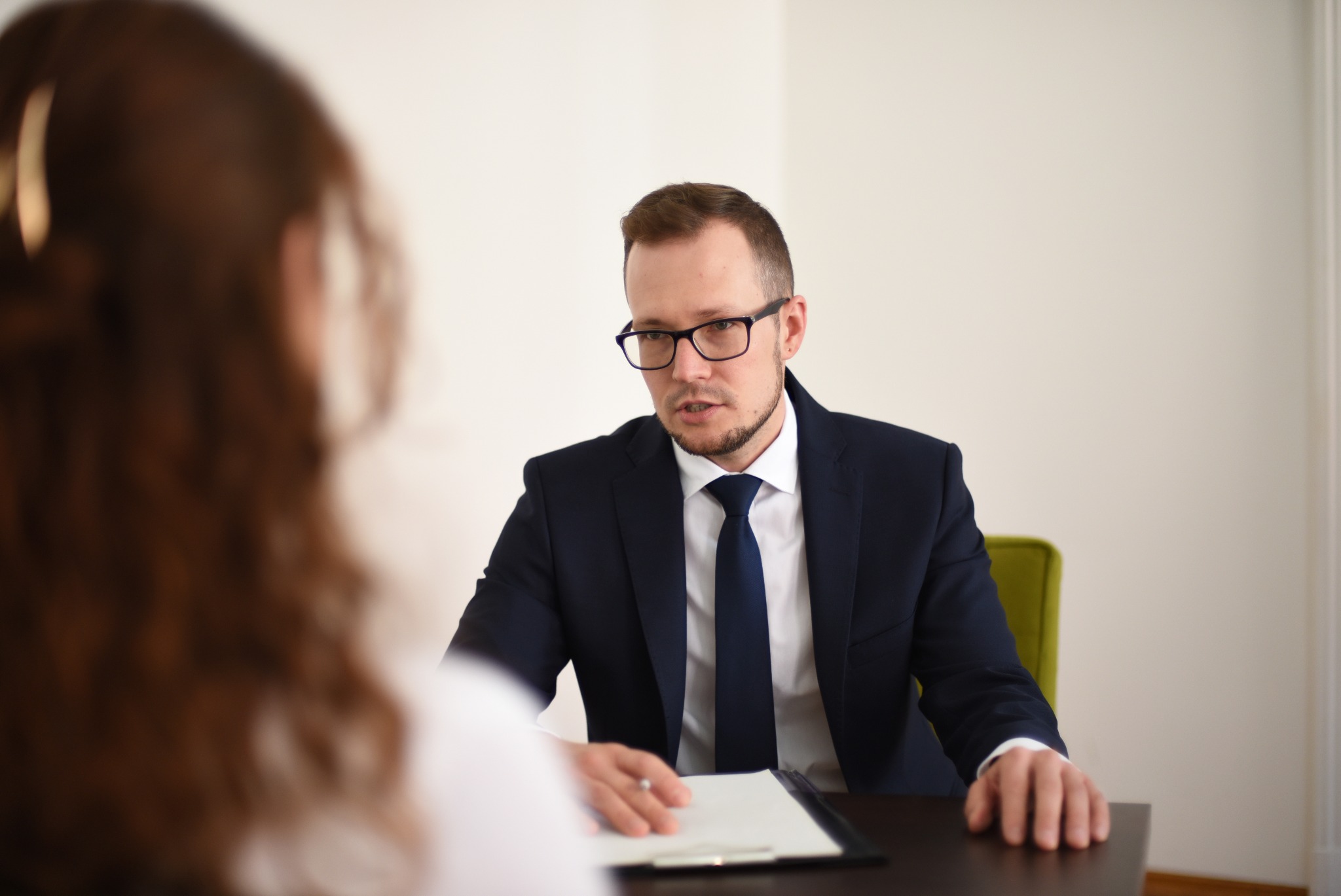 Mężczyzna w garniturze i okularach siedzi za biurkiem, prowadząc rozmowę z osobą siedzącą naprzeciwko, której widać jedynie fragment włosów i ramienia. Na biurku leży podkładka z kartkami.