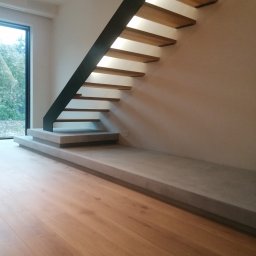 NICE NICE DESIGN - Nowoczesne schody wiszące z drewnianymi stopniami i czarną metalową konstrukcją, oświetlone od spodu, z betonową podstawą i drewnianą podłogą w minimalistycznym wnętrzu.