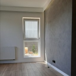 NICE NICE DESIGN - Pomalowane na szaro pomieszczenie z drewnianą podłogą, białym grzejnikiem i oknem z widokiem na zewnątrz, z detalami takimi jak listwy przypodłogowe i gniazdko elektryczne.