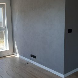 NICE NICE DESIGN - Szara ściana z efektem dekoracyjnym, listwa przypodłogowa, drewnopodobna podłoga i okno z widocznym światłem słonecznym w rogu pokoju.