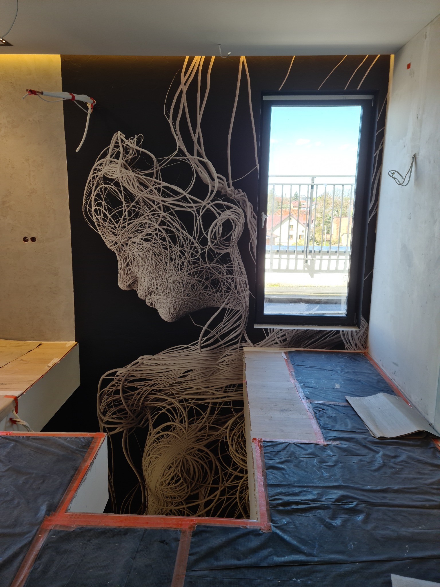 Pokój w trakcie wykańczania z czarną ścianą z grafiką przedstawiającą profil twarzy wykonaną z białych linii, podłoga zabezpieczona folią, widok na balkon przez okno.