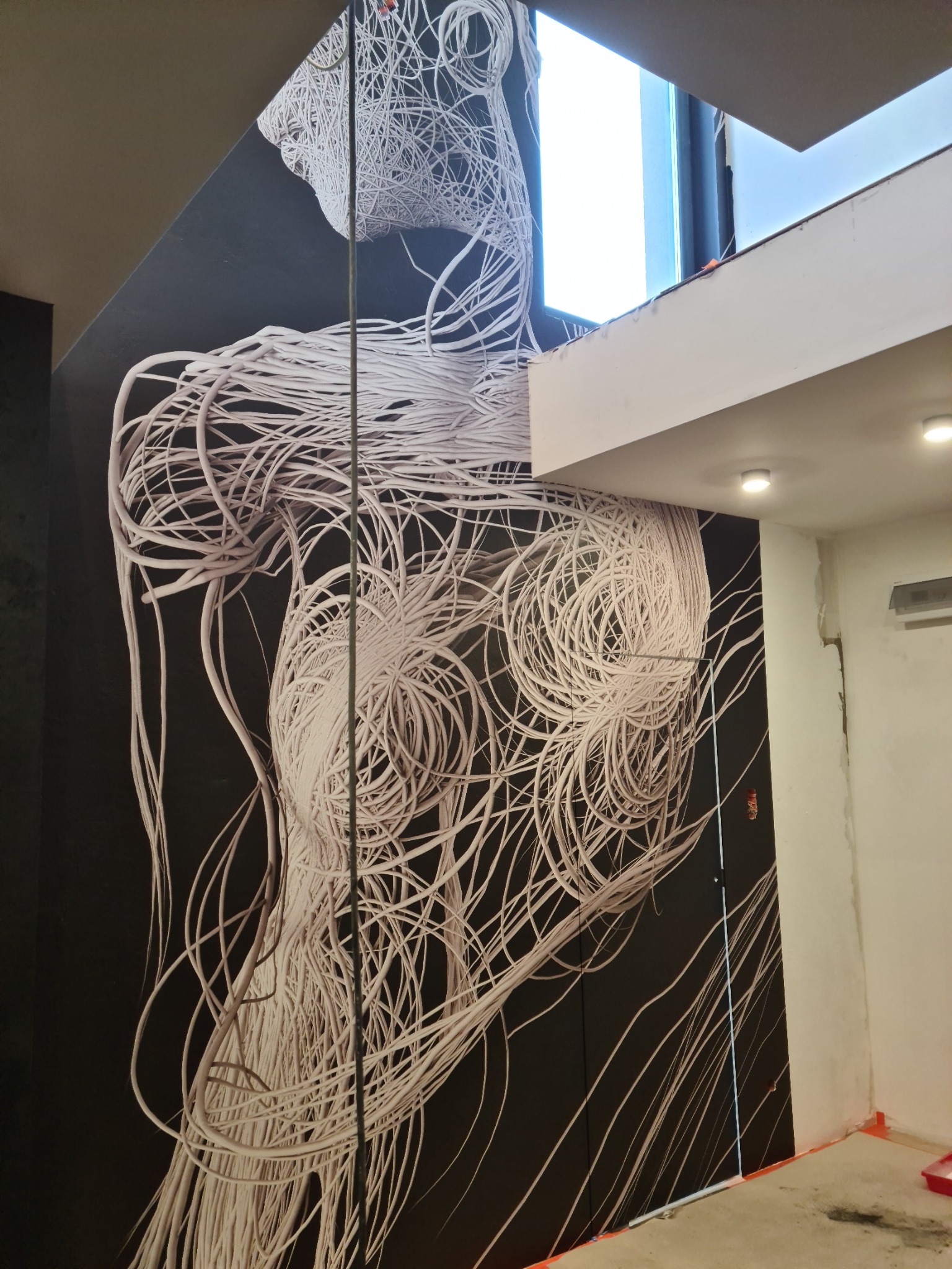 Abstrakcyjna fototapeta z motywem splątanych białych kabli na czarnym tle, widoczna na ścianie w trakcie wykańczania wnętrza, z fragmentem okna i sufitu