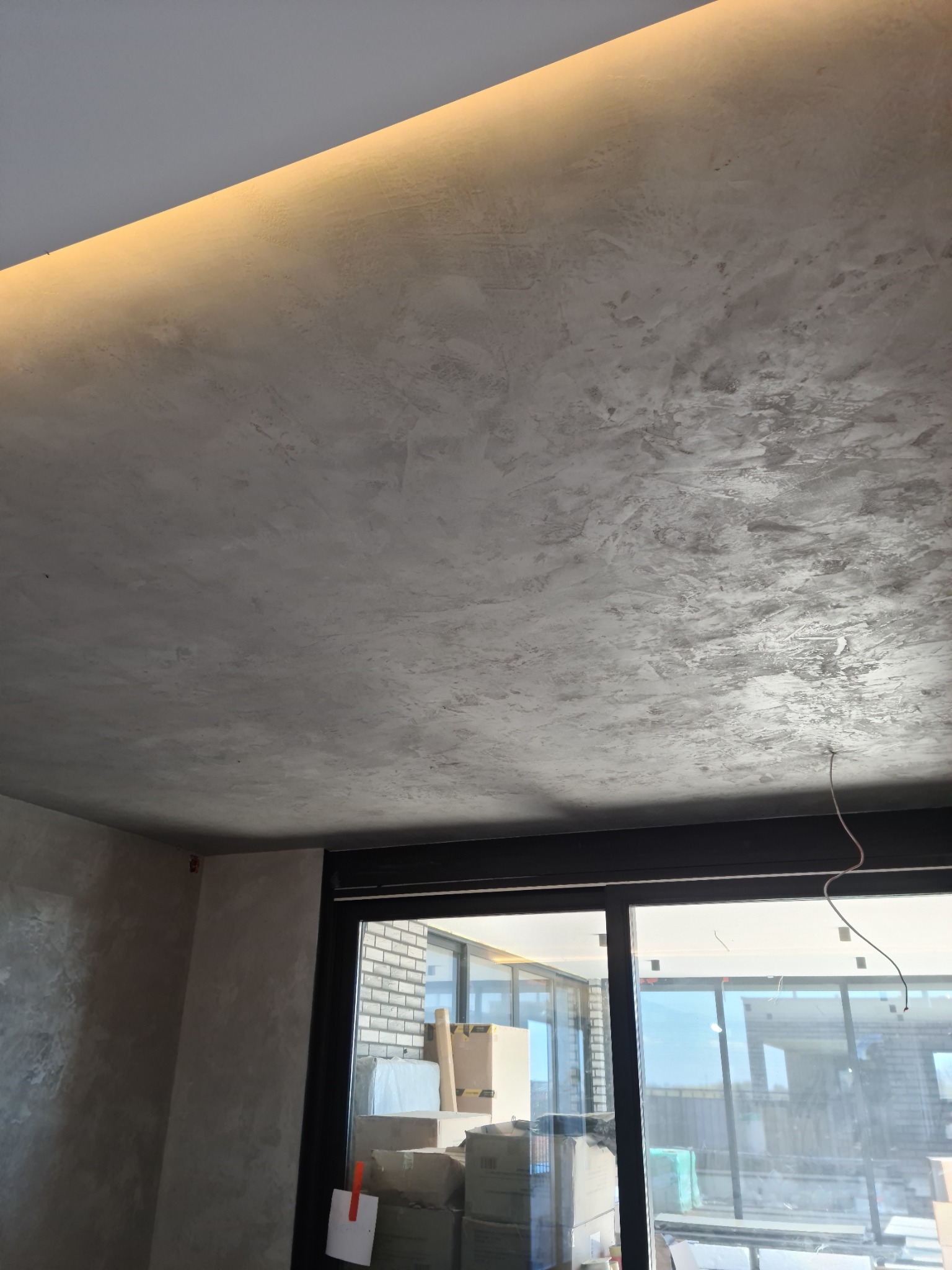 Szara, teksturowana ściana i sufit z podświetleniem LED, widoczne kable i okno z widokiem na budynek w budowie.